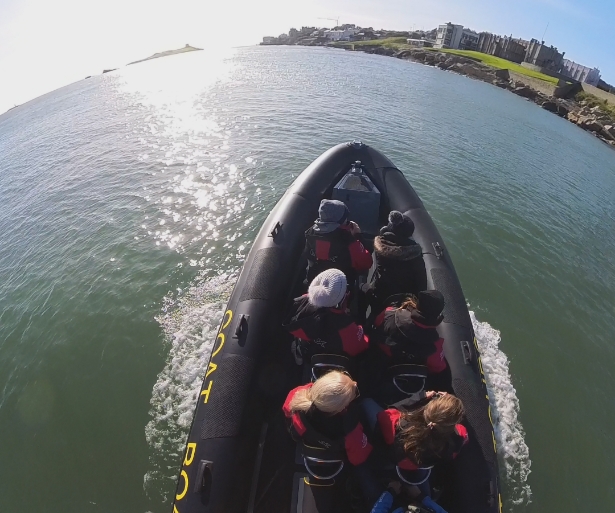 A group of people wearing life jackets are seated in a black inflatable boat. The boat is cruising on calm water under a clear sky. A distant shoreline with buildings and green grassy areas is visible ahead.