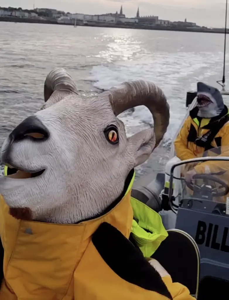 Two people in yellow jackets on a boat, one wearing a goat mask and the other a shark mask. The boat speeds on water with a city skyline in the background. The scene is playful and unconventional.