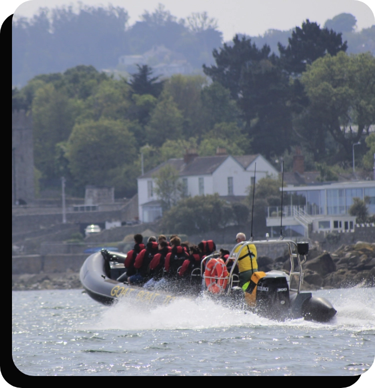 A group of people wearing red and black life jackets ride a black inflatable motorboat on a body of water. The background features lush green trees and buildings on the shoreline under a partly cloudy sky.