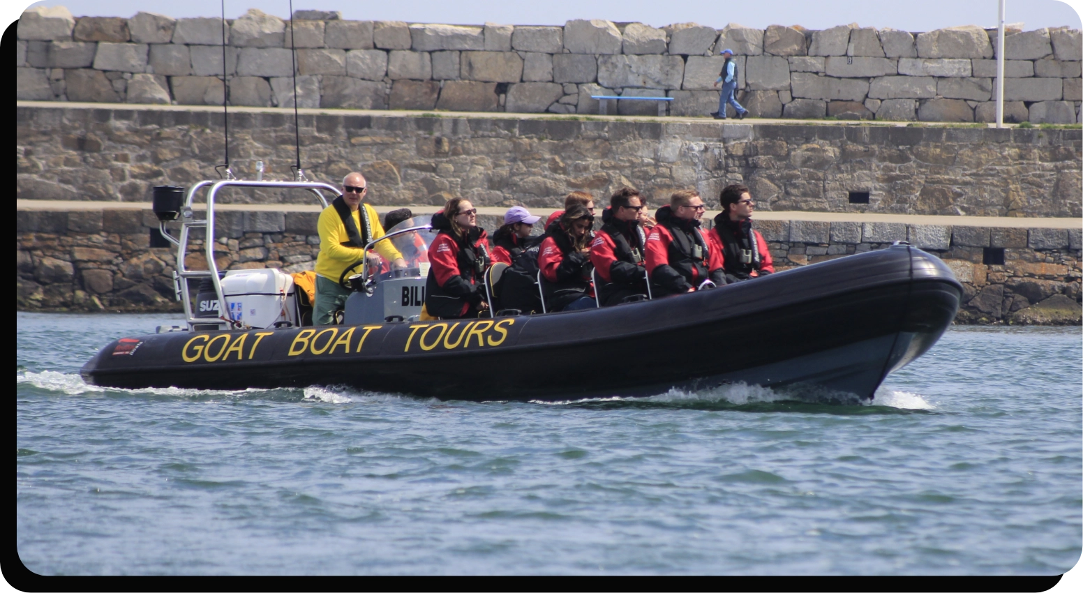 A group of people wearing life jackets are seated in a black inflatable boat labeled "GOAT BOAT TOURS" on the side. A person in a yellow jacket is steering the boat, which is moving through the water near a stone wall.