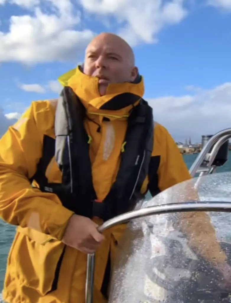 A person wearing a yellow jacket and life vest is steering a boat. They are looking into the distance with a cloudy sky and water in the background.