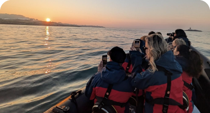 People on a boat take photos of the sunrise over the ocean. They are wearing blue and red jackets and standing close together. The sun is low on the horizon, casting a warm glow over the water.