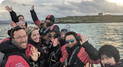 A group of people wearing red and black jackets wave while seated in a boat on the water. The background features an island with a stone tower and a cloudy sky.