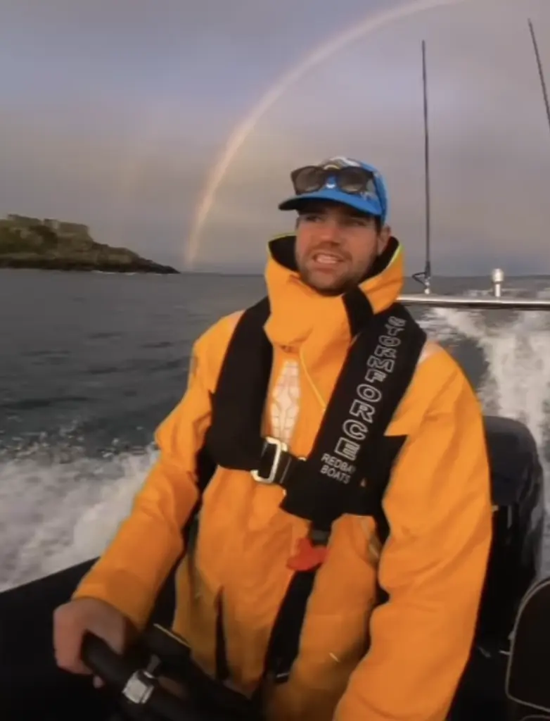 Man in a yellow raincoat operates a boat, with a rainbow arching across the cloudy sky in the background. The sea is calm, and the water is splashing around the boat. An island or small landmass is visible on the horizon.