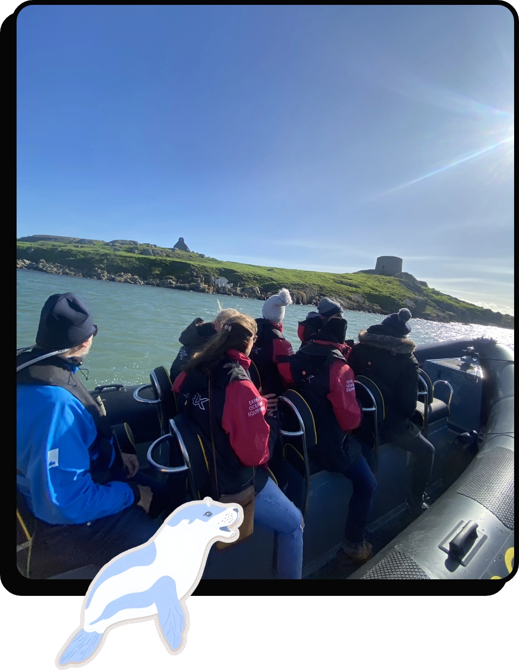 A group of people in life jackets sit in a speeding boat, looking towards an island with a stone tower. The island is covered in green grass under a clear blue sky. A cartoon seal is overlaid on the bottom of the image.