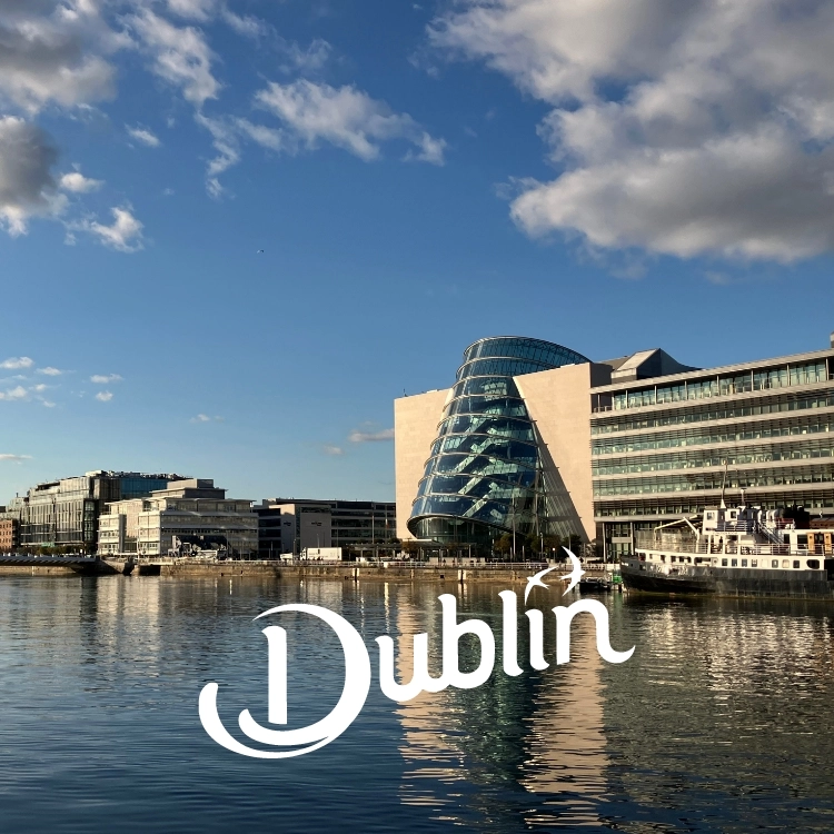 Modern waterfront buildings with a distinctive curved glass structure in the center, reflecting on the calm water. The sky is clear with a few clouds. The text "Dublin" appears artistically in the foreground.