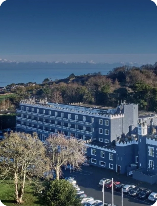 Aerial view of a large gray castle-like building with multiple windows and crenellated rooftops. The structure is surrounded by trees and a parking area with several cars. In the background, there is a vast body of water under a clear blue sky.