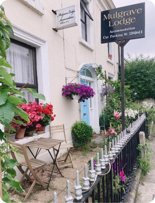 Exterior of Mulgrave Lodge, a quaint accommodation with a pale blue door, white walls, and vibrant hanging flower baskets. A small table and two chairs sit near the entrance beside a black wrought-iron fence. Sign with contact details is visible.