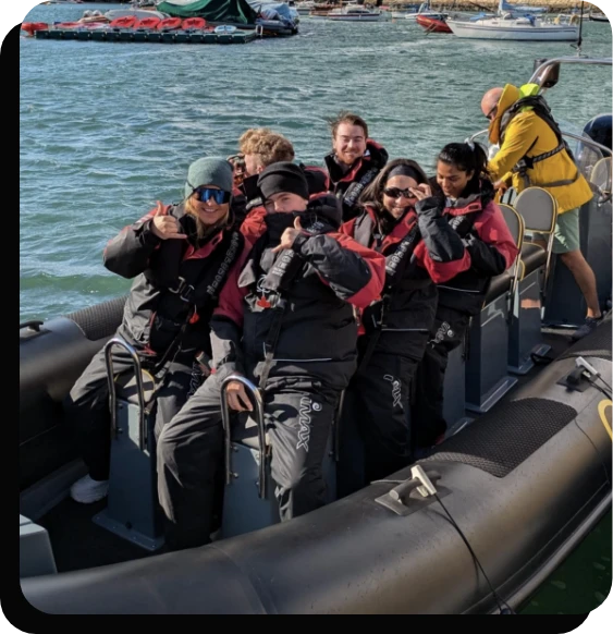 A group of people wearing black waterproof suits and red life jackets sit in a gray inflatable boat on the water. They are smiling and giving thumbs up as another person operates the boat. Other boats and a dock are visible in the background.