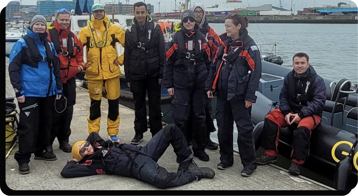 A group of nine people in sailing gear pose on a dock near a rib boat. Eight are standing or sitting, while one person lies on the ground with legs crossed. The background features a harbor with boats and industrial buildings.