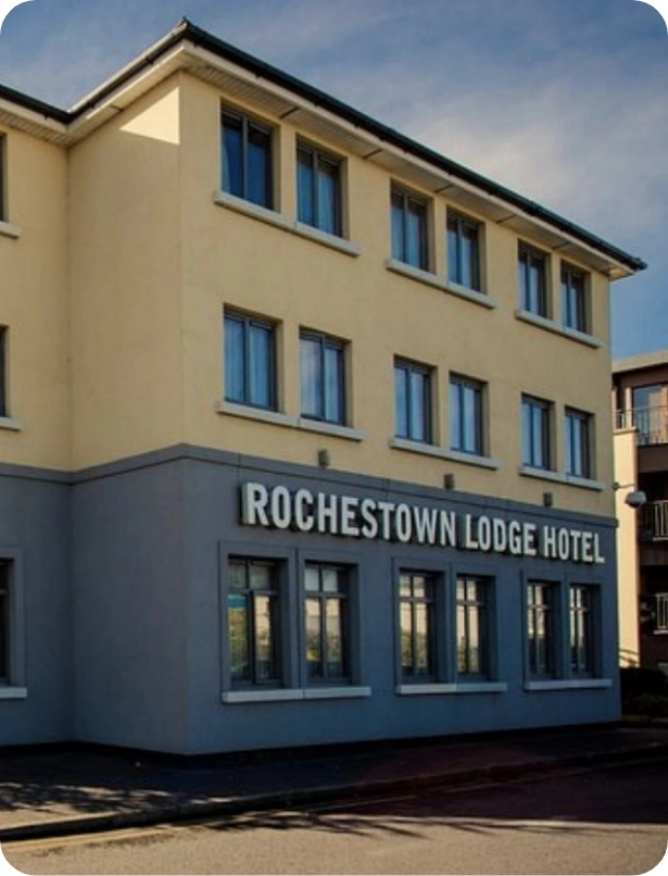 Street view of a three-story beige building with "Rochestown Lodge Hotel" signage. The building features multiple windows and a partial glimpse of another structure to the right. The sky is clear and blue.