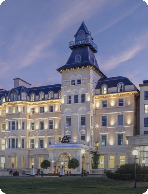 A grand, multi-story hotel with ornate architecture is illuminated against a twilight sky. It features a central tower with a "Hotel" sign, numerous windows, and a small entrance canopy. The scene is tranquil and inviting.