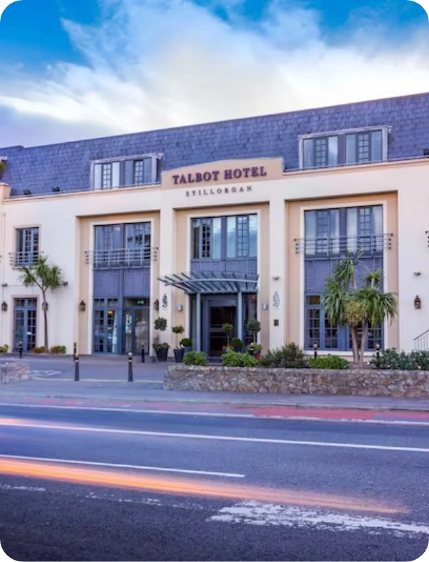 Front view of Talbot Hotel in Stillorgan, featuring a modern three-story building with large windows and a dark roof. The entrance is covered by a glass canopy, and palm trees are planted nearby. A road with light trails is in the foreground.